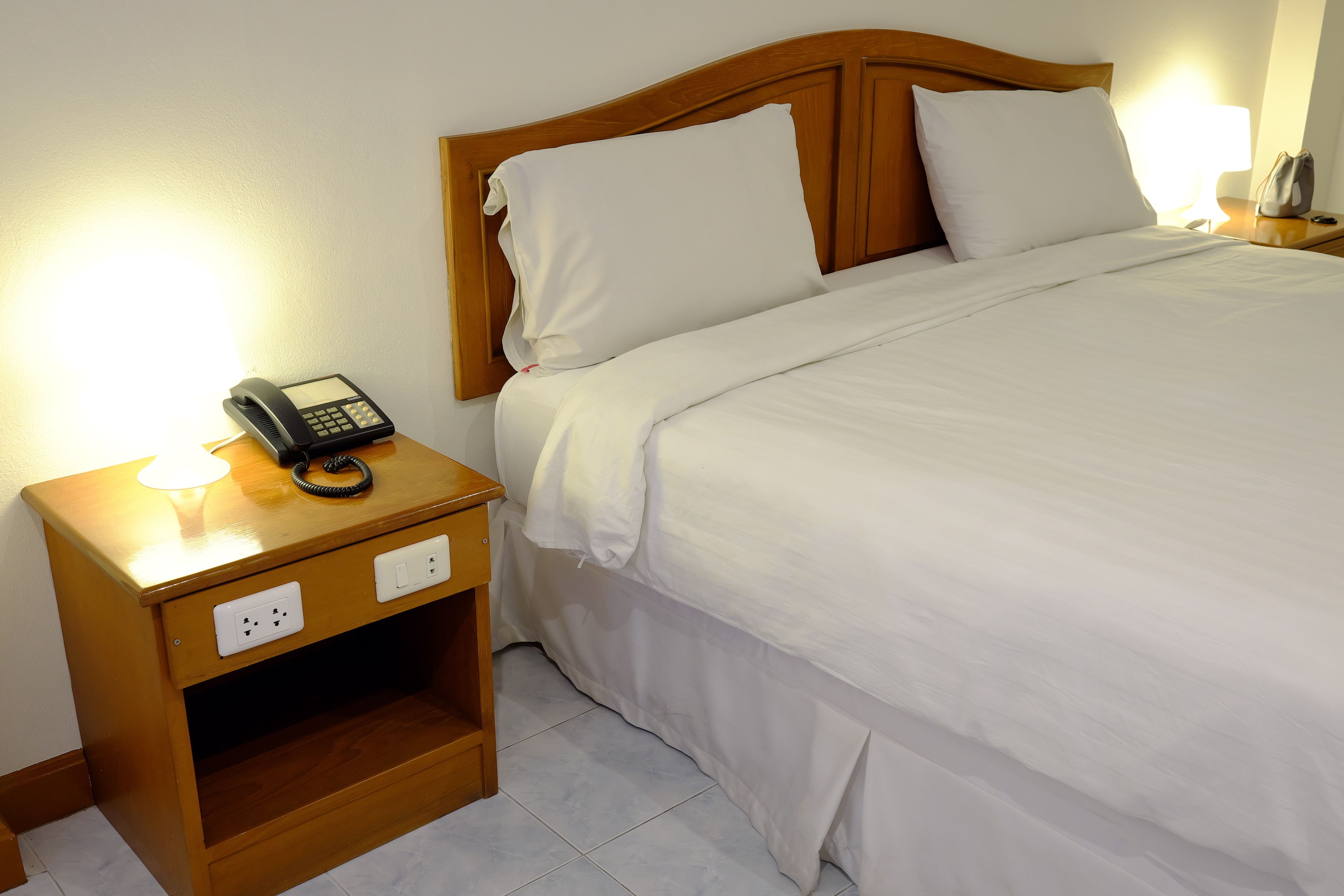 A neatly made bed with white linens, a wooden headboard, and a bedside table with a telephone and a lamp.