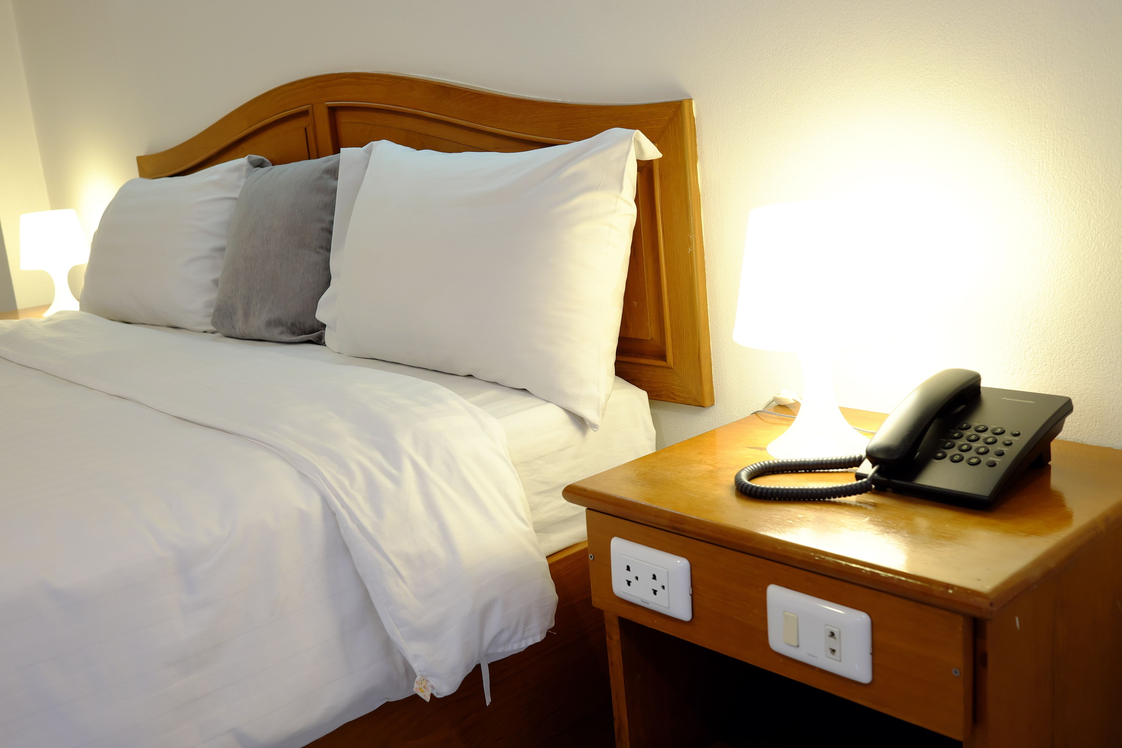 A neatly made bed with white and gray pillows, a wooden headboard, and a bedside table with a phone and a lamp.