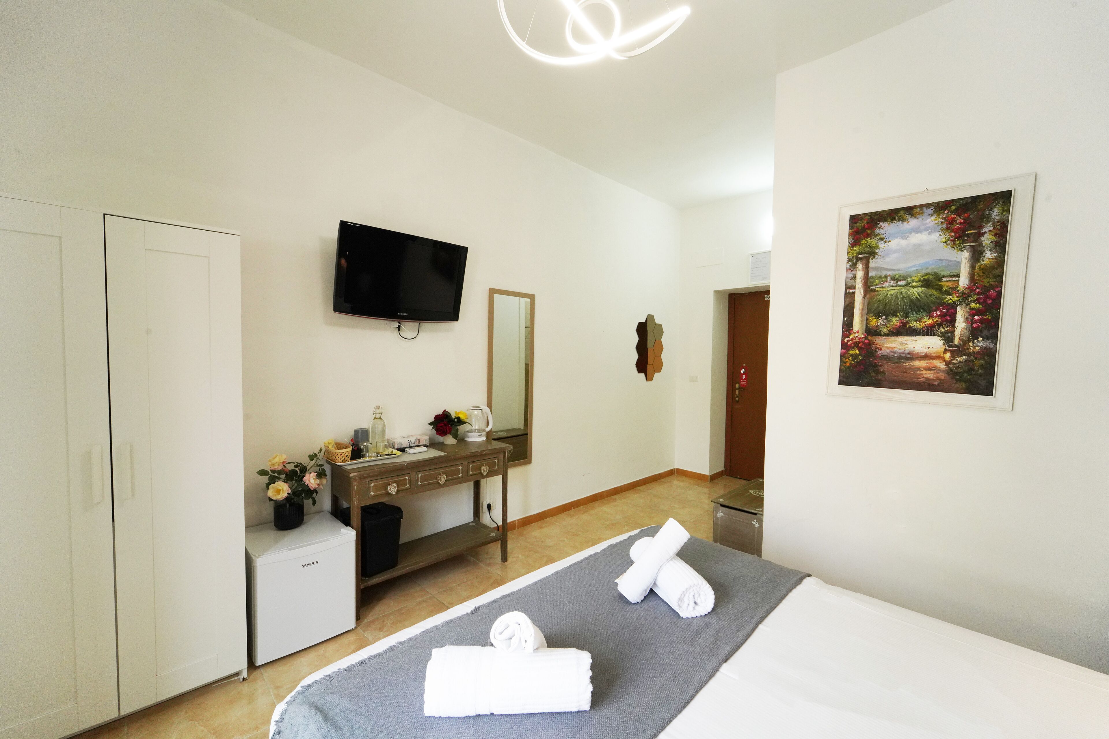 A hotel room with a bed, a TV, a desk with a mirror, a small fridge, a painting, and a wardrobe.