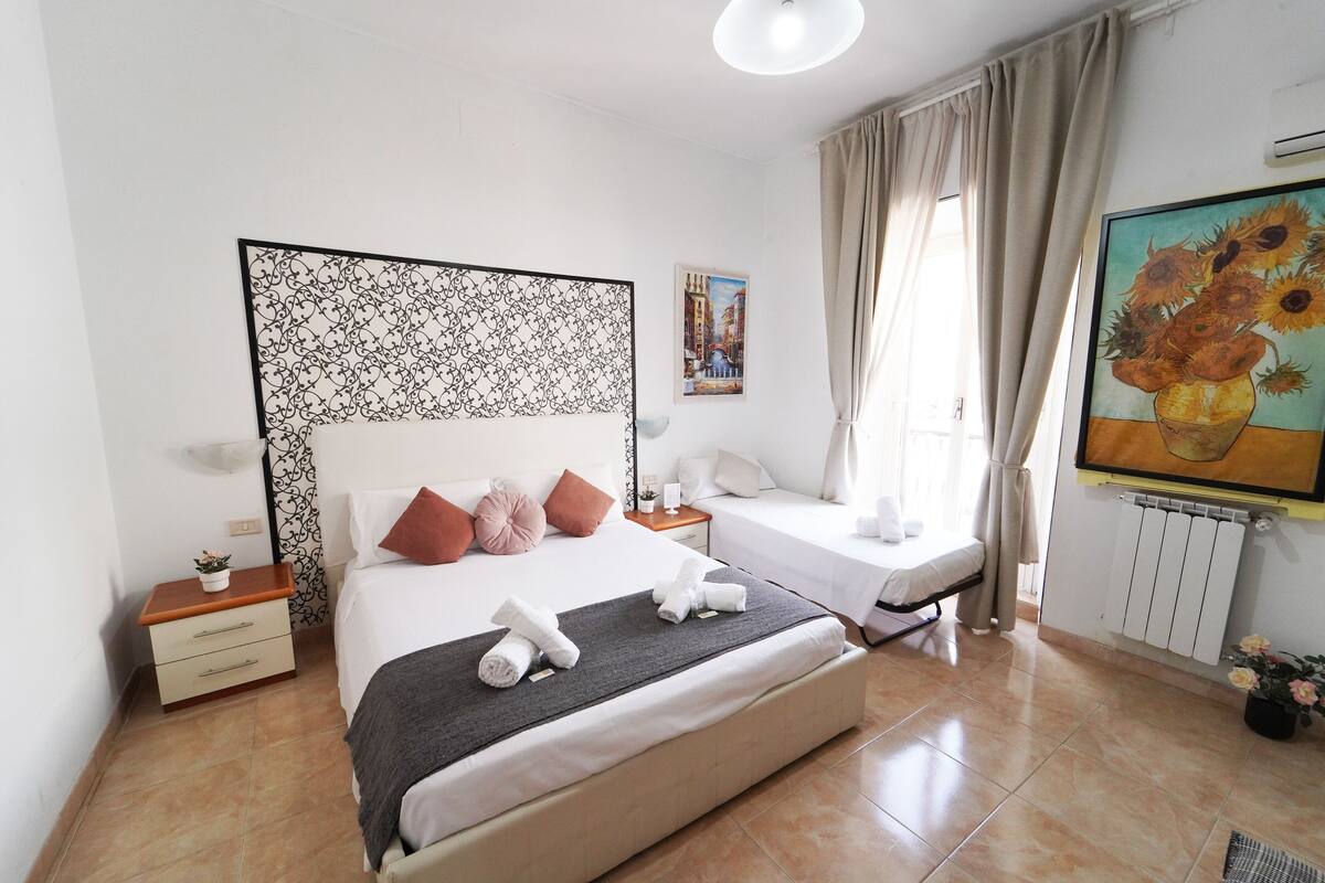 A bedroom with a large bed, a bedside table, a nightstand, a window with curtains, and a painting on the wall.