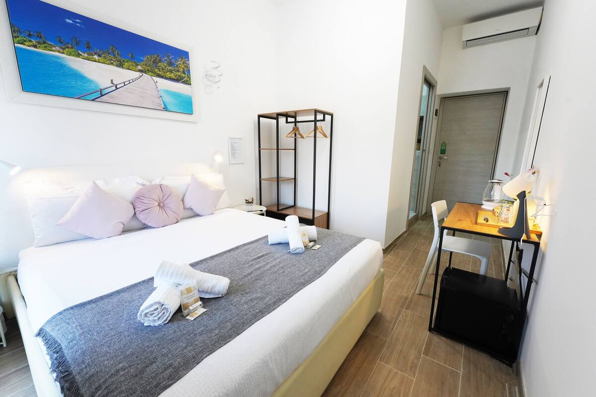 A hotel room with a bed, a desk, a chair, a wardrobe, and a painting of a beach scene.