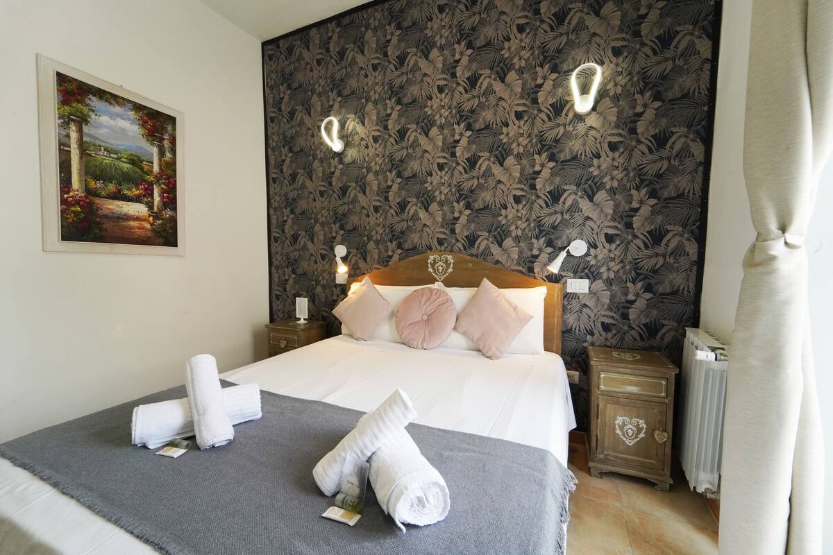A neatly made bed with a grey bedspread, white linens, and a rolled towel on the side. There is a painting on the wall and a patterned wallpaper behind the bed.