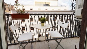 Un balcón con dos sillas blancas, una mesa redonda pequeña, una planta en maceta y vistas a un edificio blanco.
