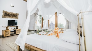 Un dormitorio con cama con dosel, un televisor sobre un pedestal de madera y una ventana con vistas a la vegetación.