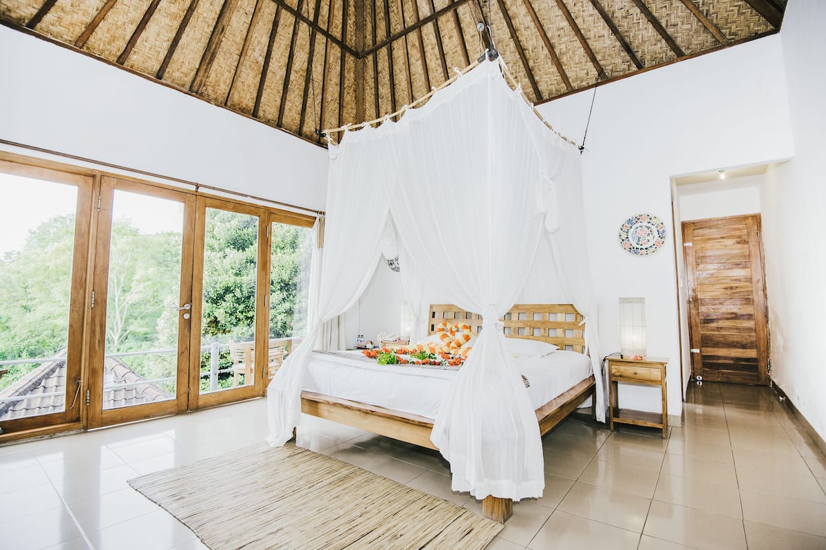 A bedroom with a four-poster bed, a wooden headboard, a nightstand, and a large window with a view of greenery.