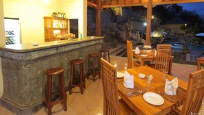 Un área de comedor al aire libre con barra, mobiliario de madera y vistas a una piscina al atardecer.