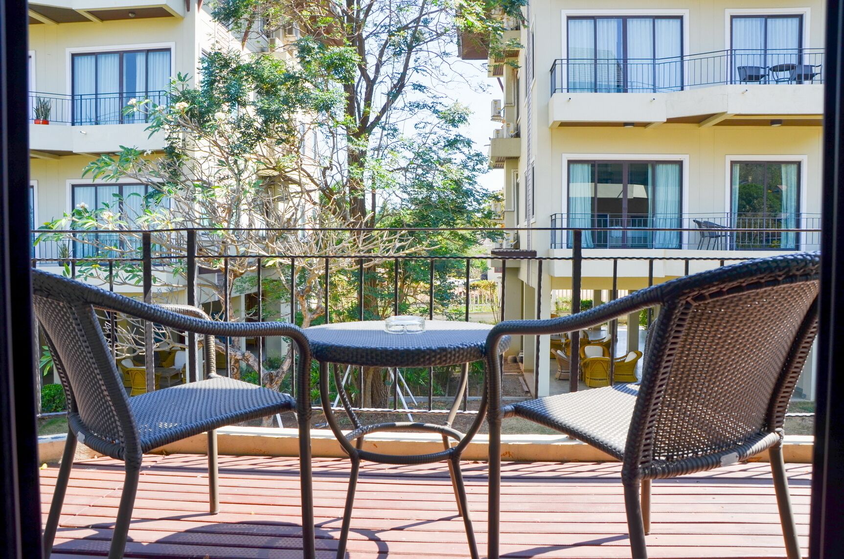 A balcony with wicker chairs and a table, overlooking a building with multiple balconies and trees.