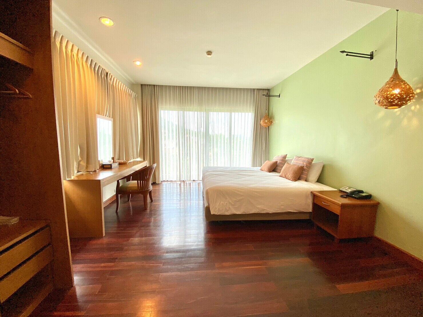 A hotel room with a wooden floor, a bed with white bedding, a desk with a chair, and a large window with curtains.