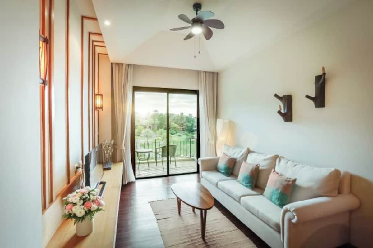A modern living room with a sofa, a coffee table, a ceiling fan, a large window with curtains, and a view of greenery outside.