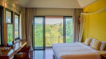 A hotel room with a large bed, a desk, and a chair. There is a balcony with a view of greenery and mountains.