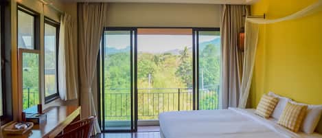 A hotel room with a large bed, a desk, and a chair. There is a balcony with a view of greenery and mountains.