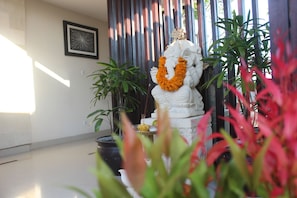 Una estatua de una deidad con una guirnalda, rodeada de plantas en macetas y un cuadro en la pared.
