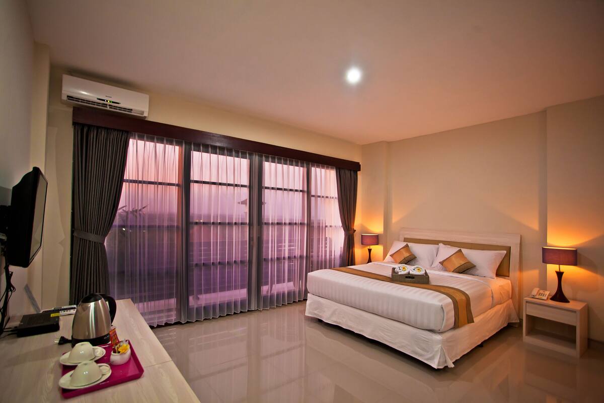 A hotel room with a bed, bedside lamps, a desk with a kettle and teapot, and a view through large windows.