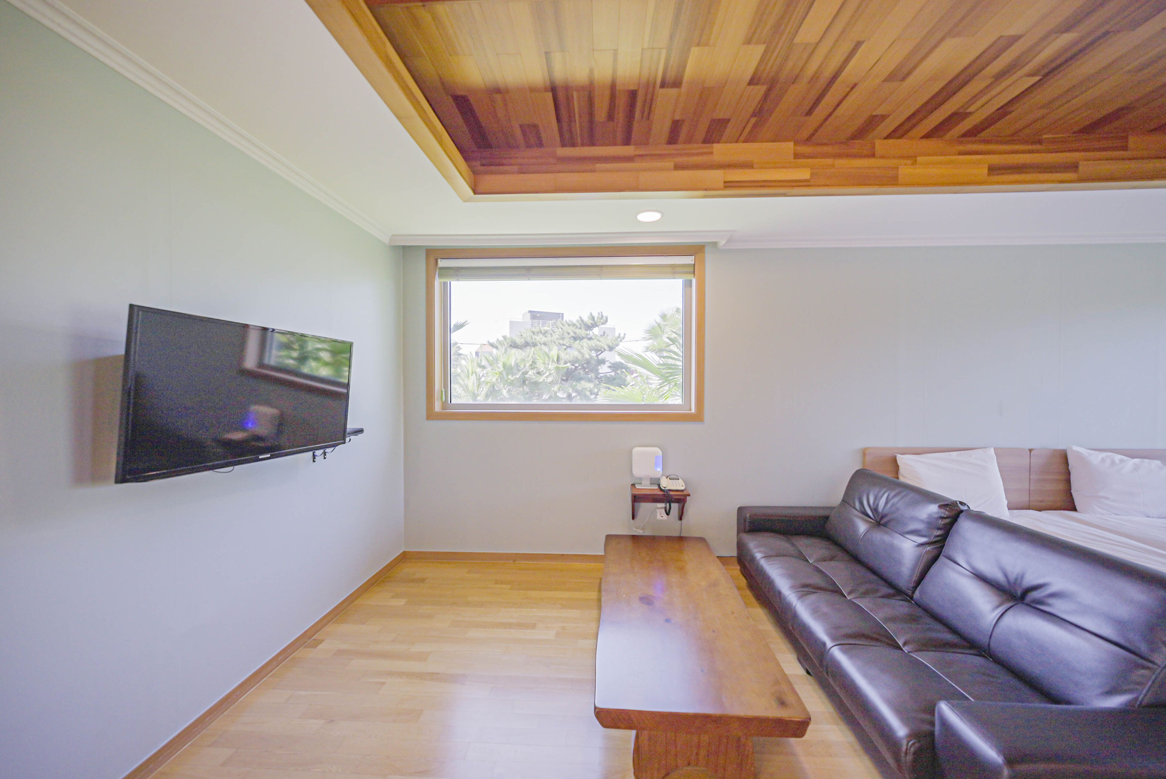 A room with a wooden ceiling, a television mounted on the wall, a window with a view of trees, a brown leather sofa, and a wooden table.