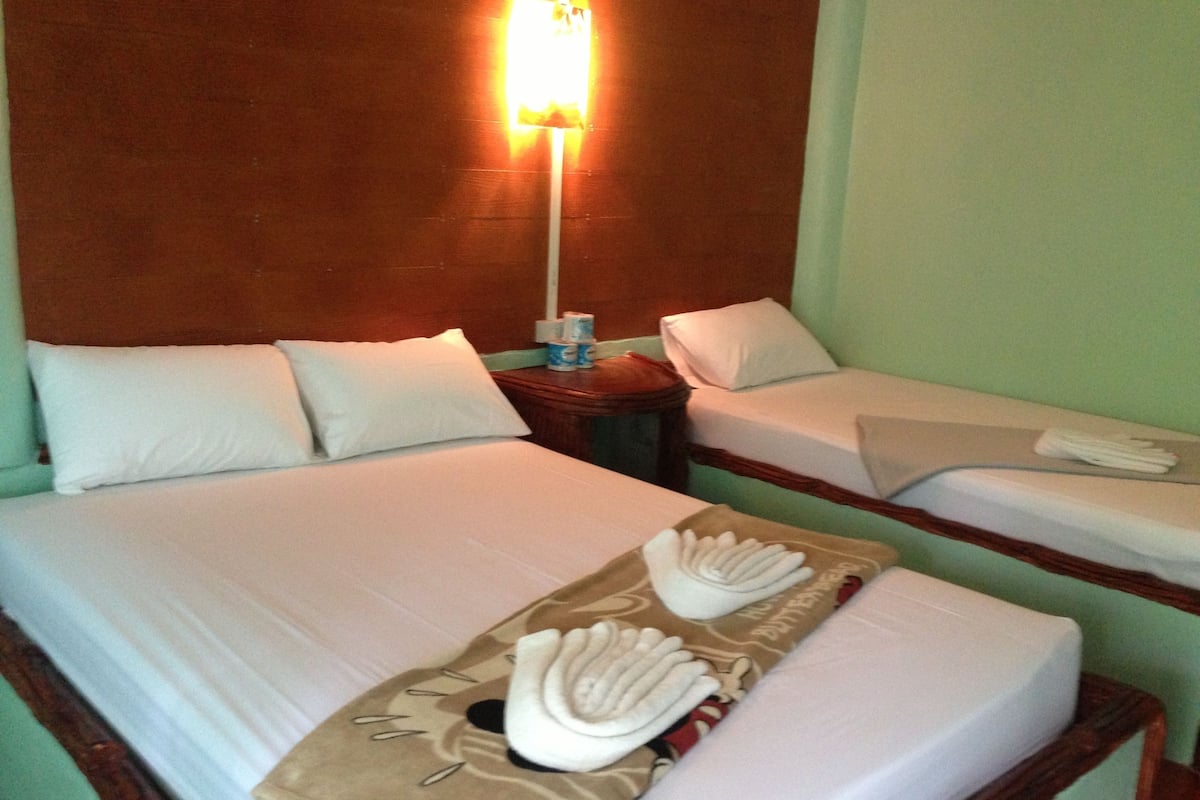 Quarto de hotel com duas camas, cada uma com roupa de cama e almofadas brancas. Apresenta uma mesa de cabeceira com uma luminária e uma pequena mesa com um estojo de lenços e uma garrafa.