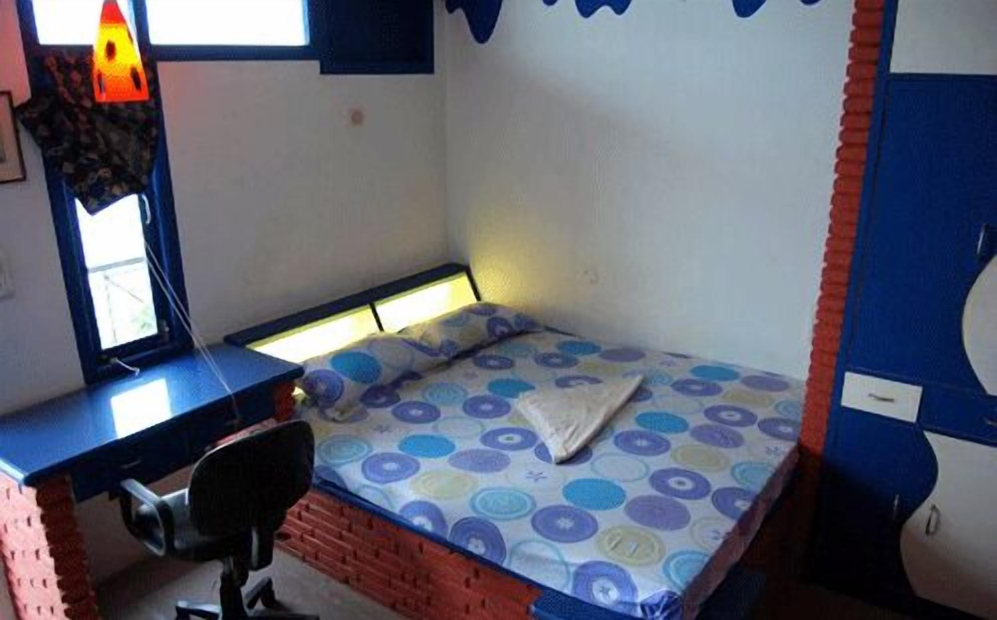 Une petite chambre avec un lit simple, un bureau et une chaise, une fenêtre et une couvre-lit à motifs.