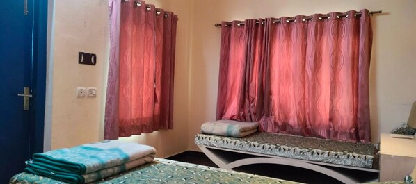 A room with two beds, each with a pillow and a folded blanket. There are curtains on the windows, and a door is visible on the left. - Rahul Guest House (Varanasi)