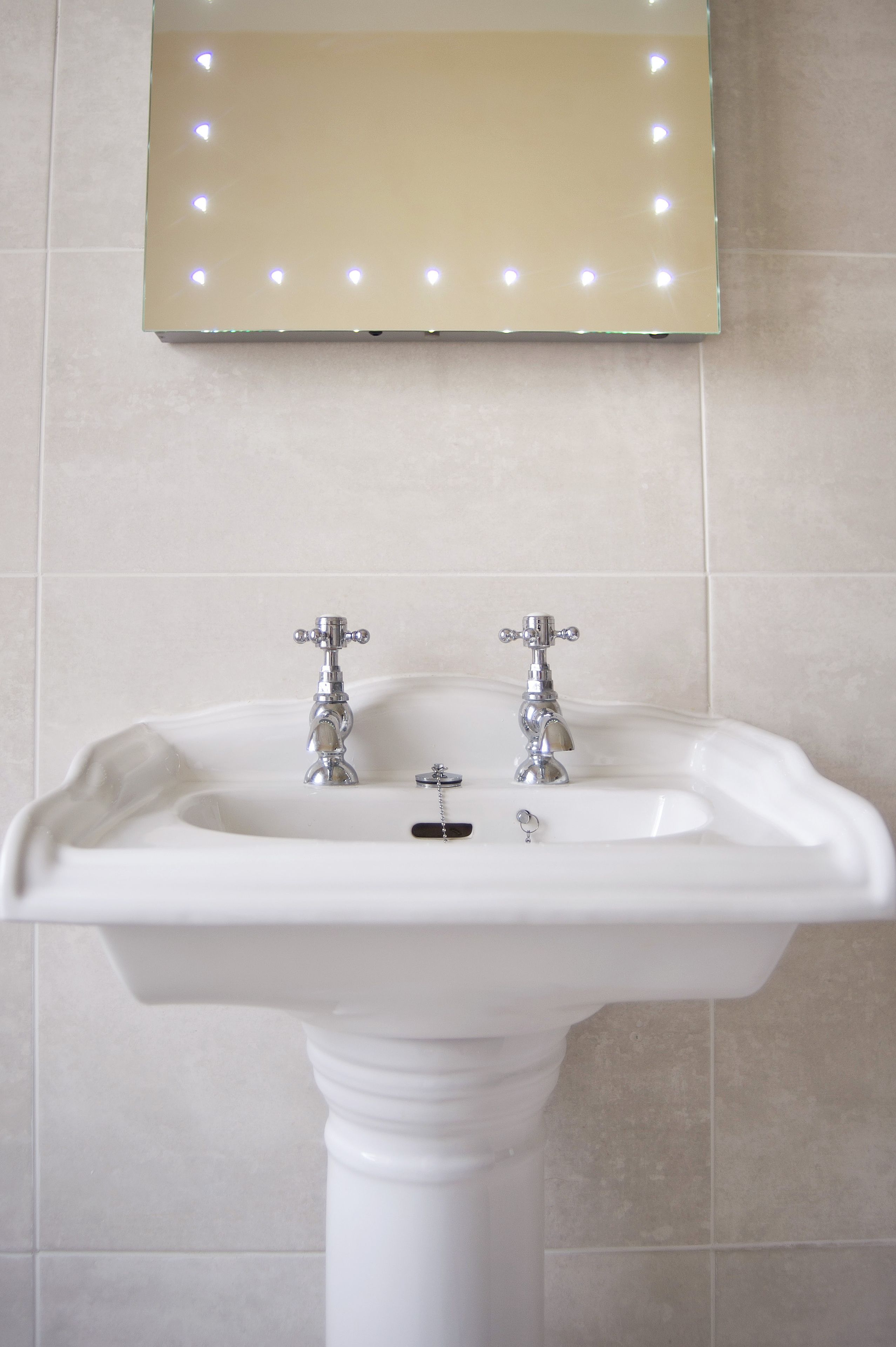 A bathroom with a pedestal sink, two faucets, and a mirror with lights.