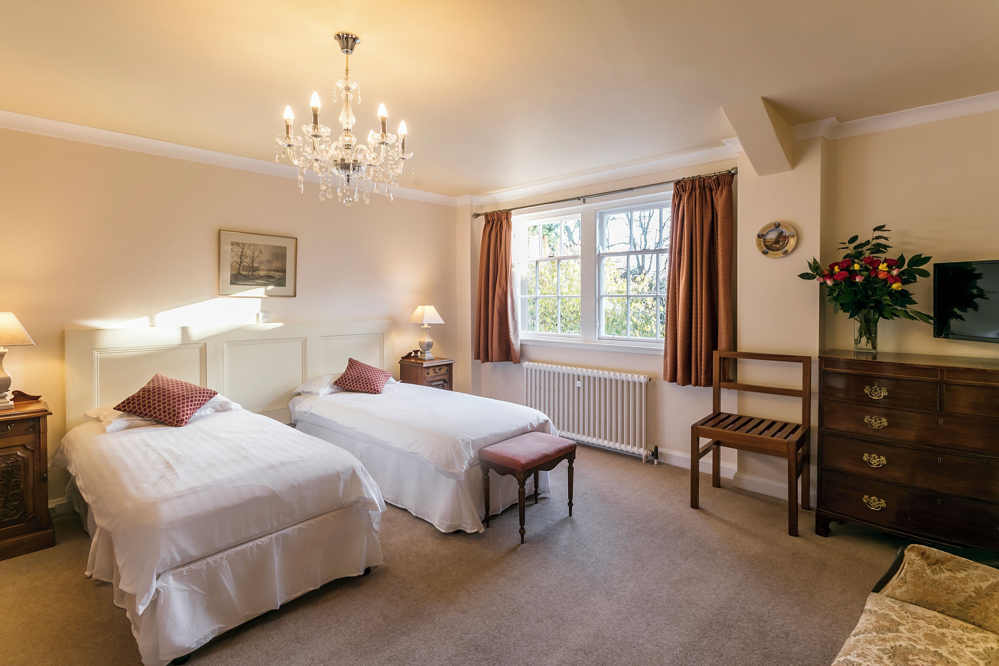 A hotel room with two single beds, a wooden dresser, a chair, a radiator, and a window with curtains.