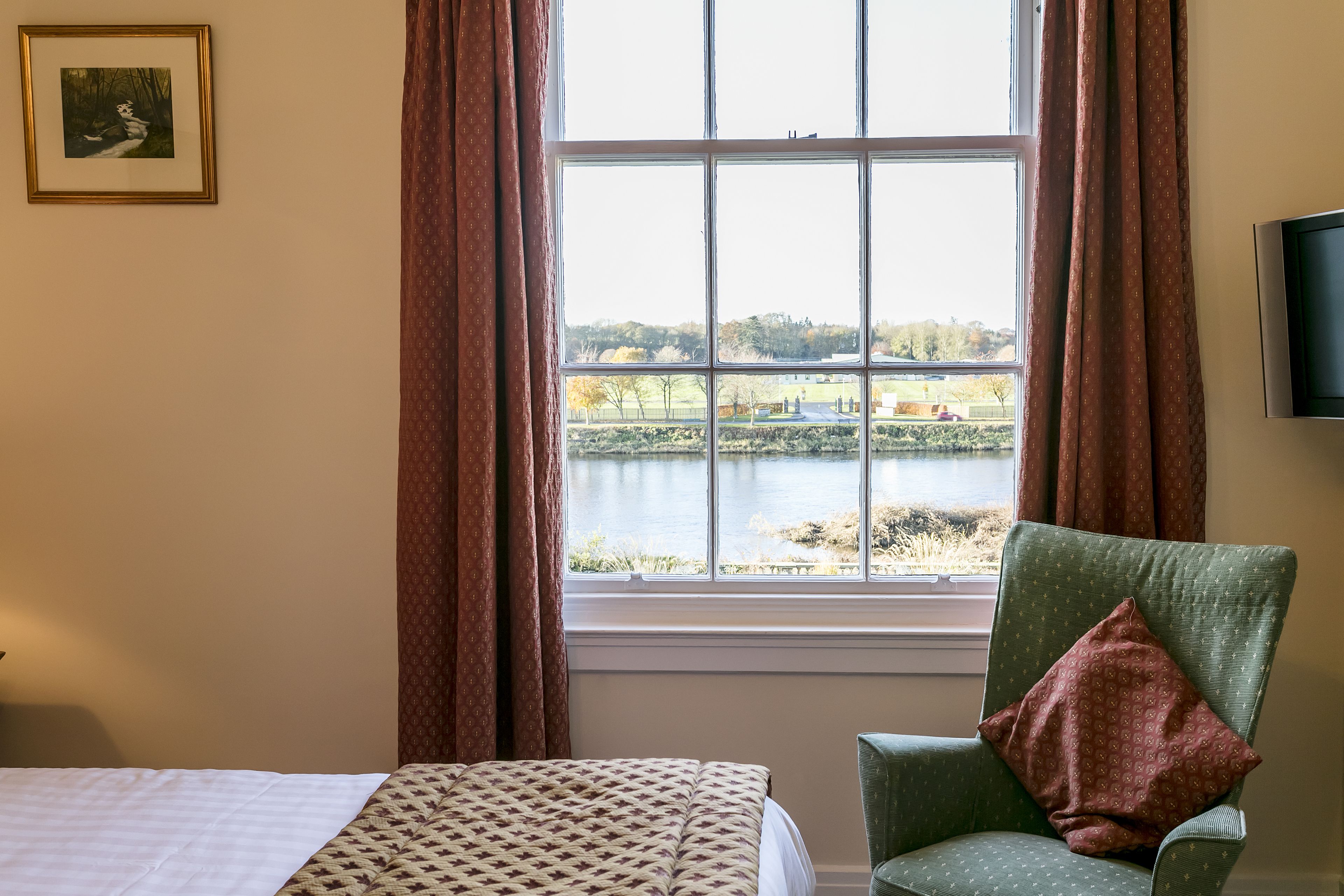A hotel room with a bed, a green armchair with a patterned pillow, a framed picture on the wall, and a window with a view of a lake and trees.