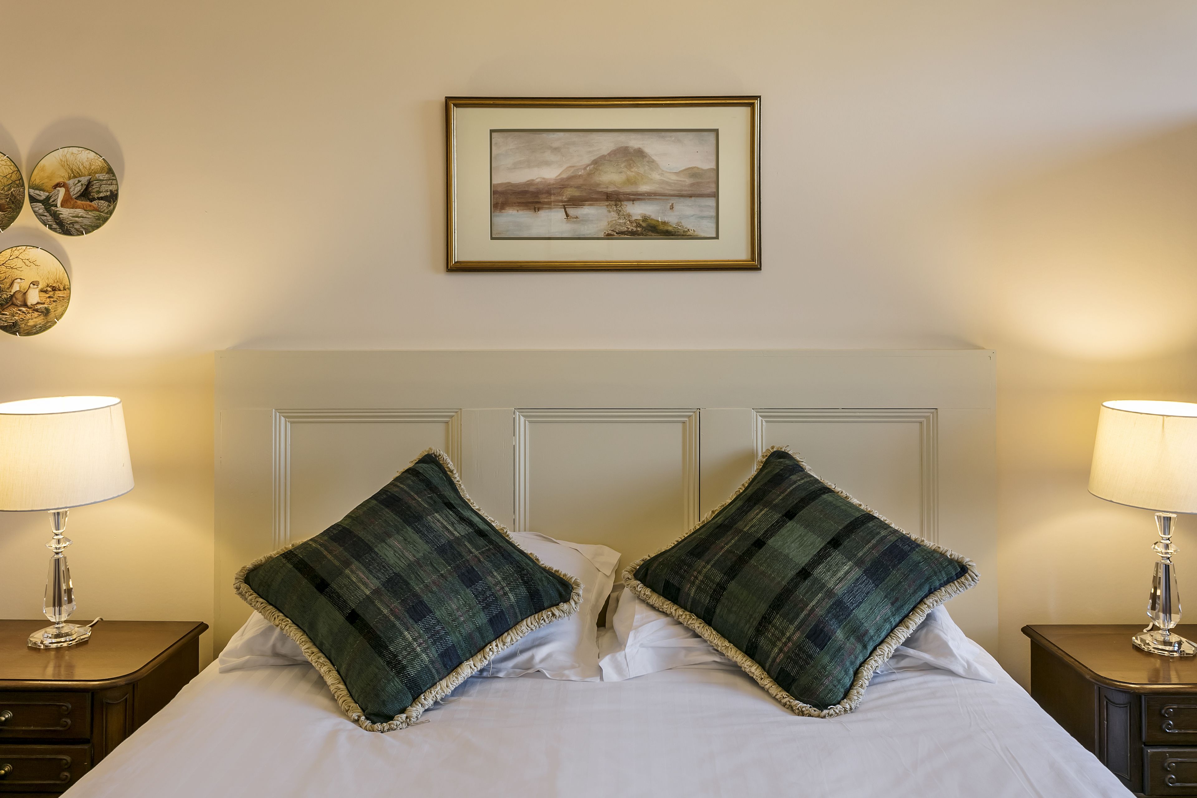 A neatly made bed with two plaid pillows, a framed landscape painting above it, and two bedside lamps on wooden nightstands.