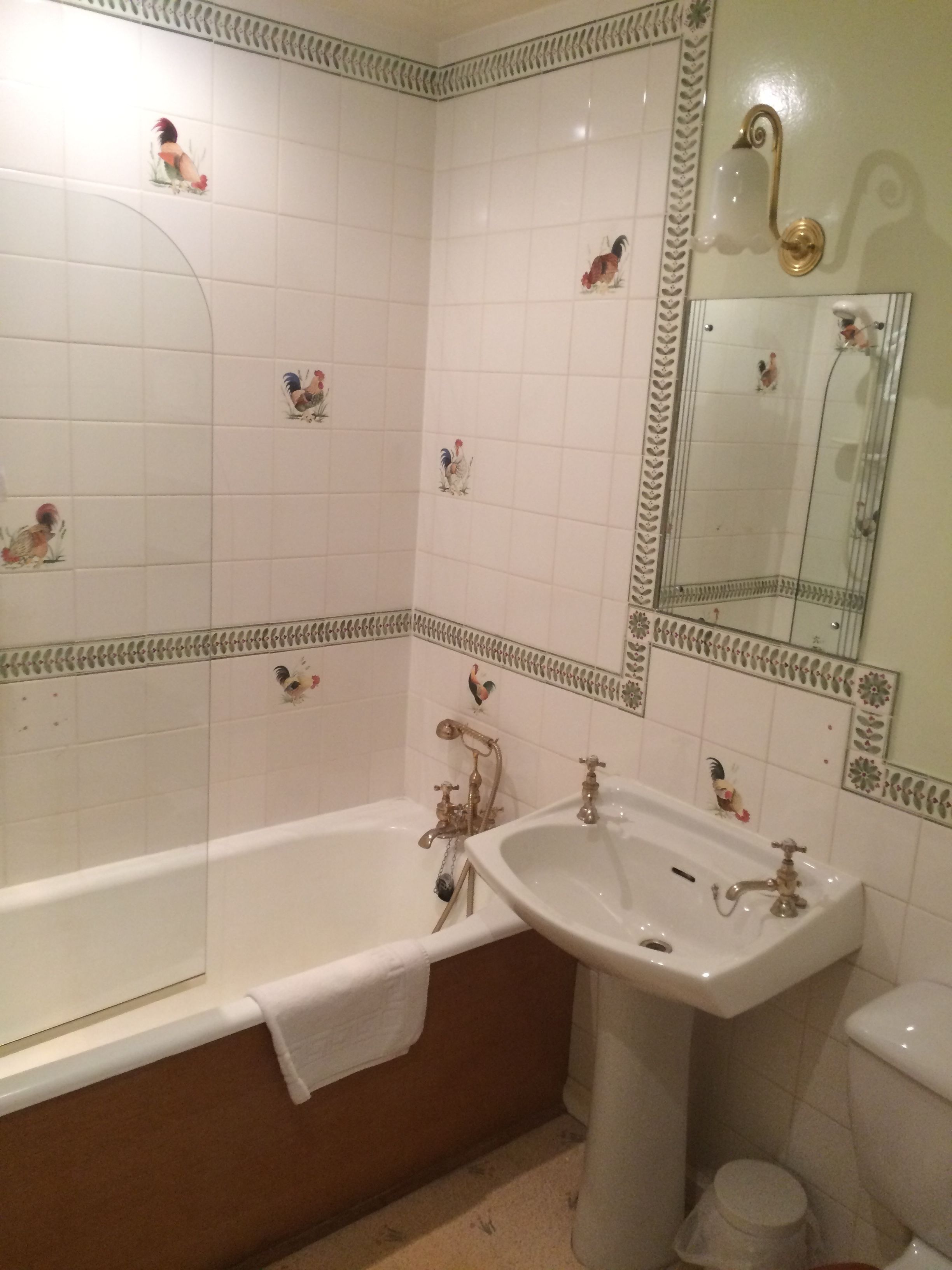 Une salle de bain avec des murs recouverts de carreaux blancs, une baignoire, un lavabo et des toilettes. On peut voir des carreaux décoratifs représentant des coqs sur les murs.