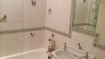 Une salle de bain avec des murs recouverts de carreaux blancs, une baignoire, un lavabo et des toilettes. On peut voir des carreaux décoratifs représentant des coqs sur les murs.