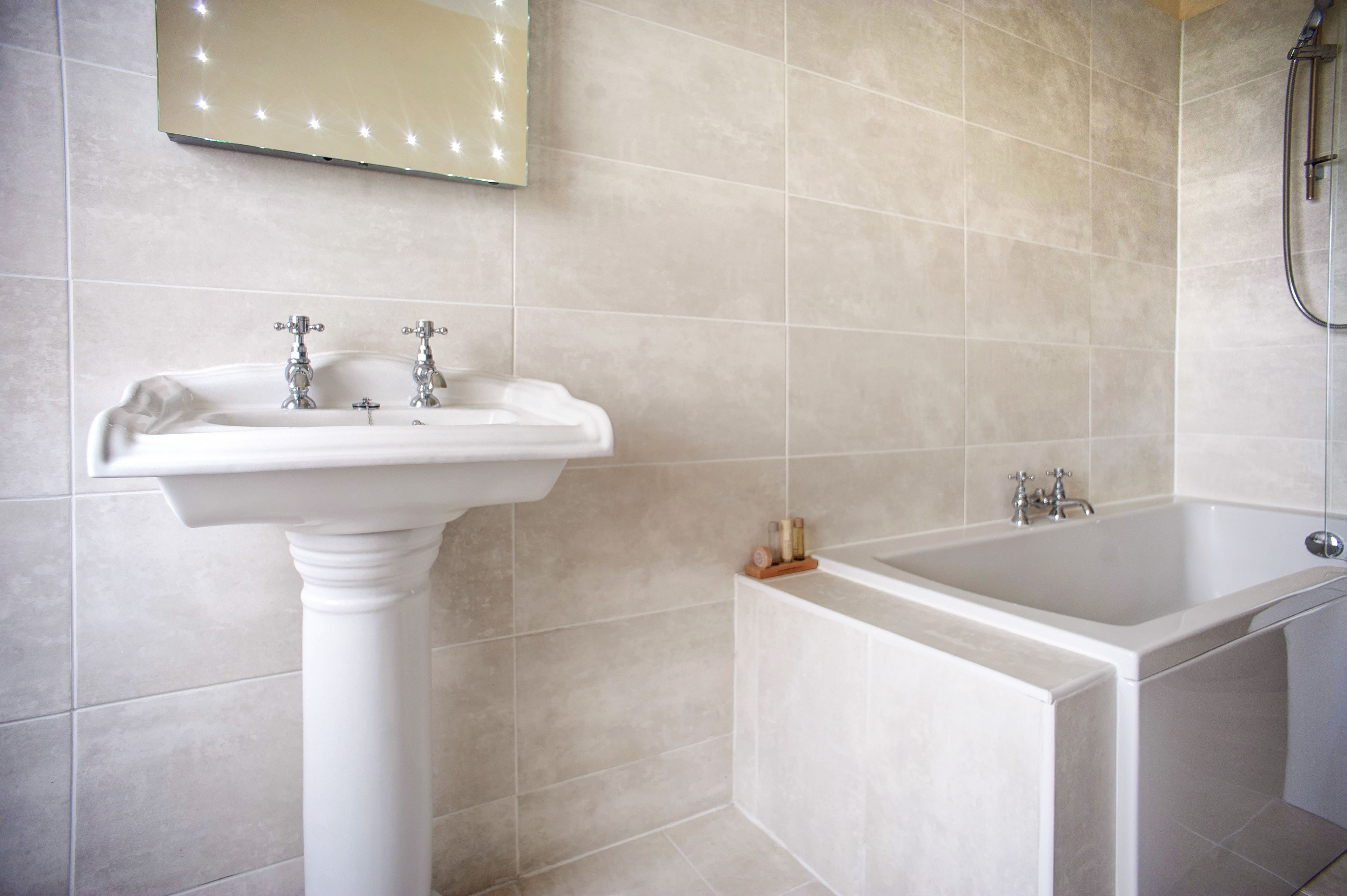 Un baño con lavamanos pedestal, bañera y espejo con luces.