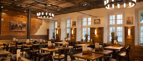 Een restaurant met houten tafels en stoelen, een bakstenen muur met een muurschildering en grote ramen met natuurlijk licht.