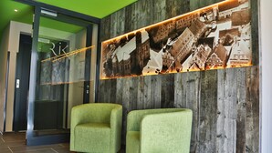 A reception area with two green armchairs, a wooden wall with a collage of historical photos, and a glass elevator door. - RiKu HOTEL Weissenhorn (Weissenhorn)