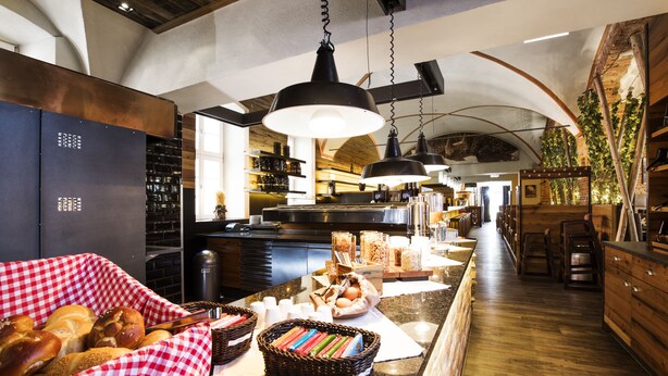 A bakery counter with bread and pastries, a checkered tablecloth, and hanging black pendant lights. - RiKu HOTEL Weissenhorn (Weissenhorn)