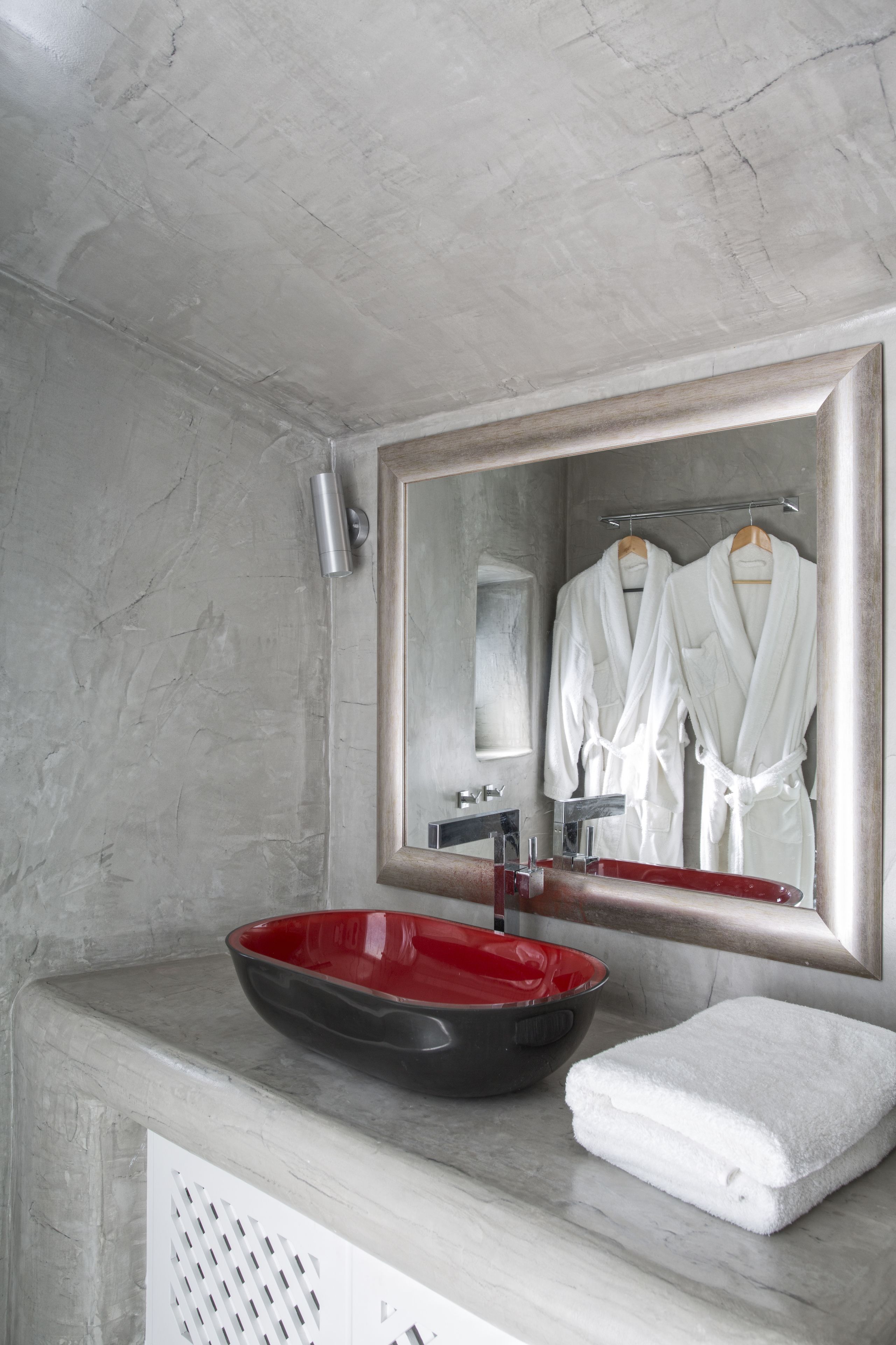 Une salle de bain avec un comptoir en marbre, un lavabo rouge, une serviette blanche et un miroir qui reflète deux peignoirs blancs.