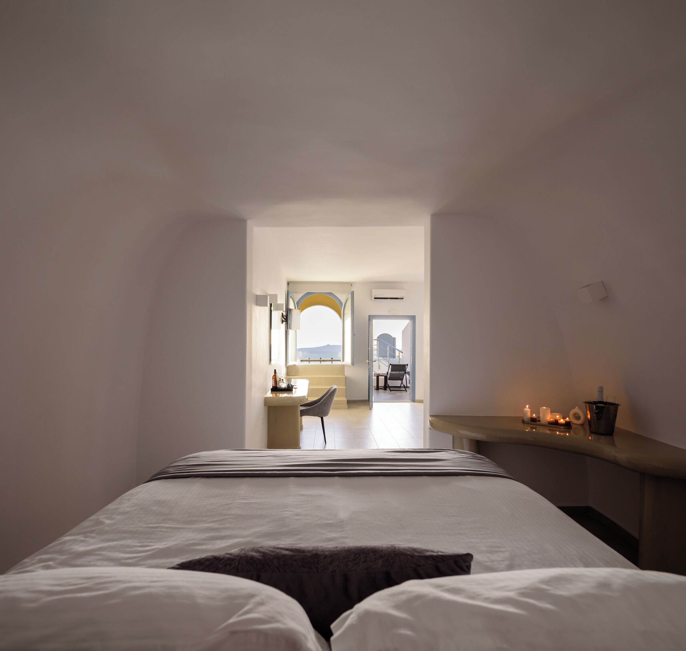 Quarto de hotel com uma cama grande, uma escrivaninha com cadeira e vista da cidade através de uma porta em arco.