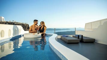 Un couple est assis au bord d’une piscine, avec une vue sur la mer et un bâtiment blanc en arrière-plan.