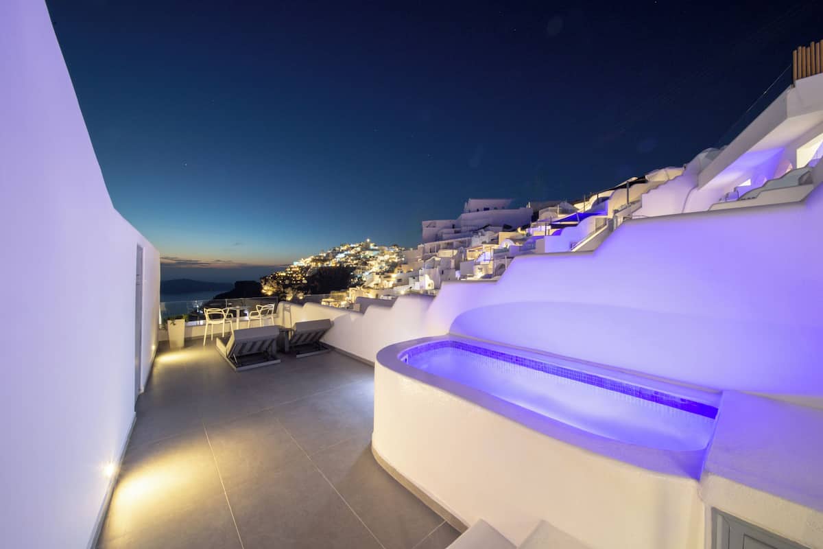 A rooftop terrace with a pool, seating area, and a view of a hillside town at dusk.