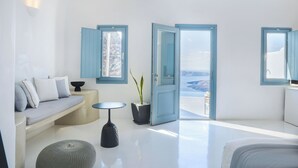 A modern, minimalist room with a sofa, a small table, and a potted plant. There are two doors leading to an outdoor area with a view. - Pegasus Suites & Spa (Santorini)