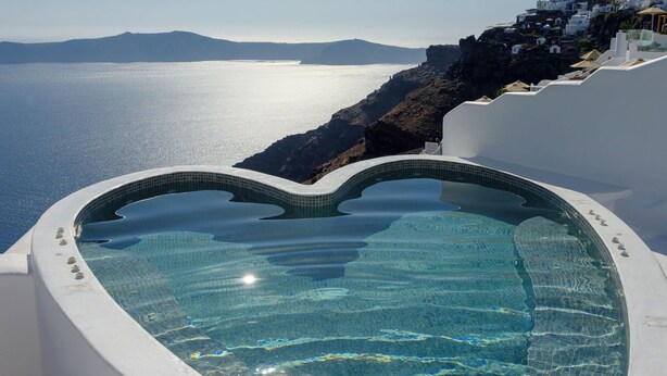 A heart-shaped infinity pool with clear blue water, overlooking a coastal landscape with white buildings and a view of the sea. - Pegasus Suites & Spa (Santorini)