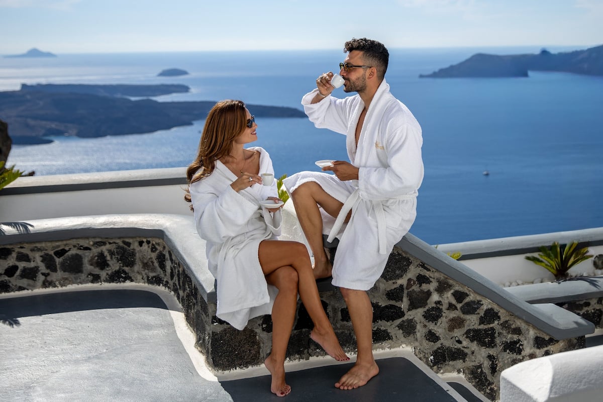 Two people in white bathrobes sitting on a stone bench overlooking a scenic ocean view.