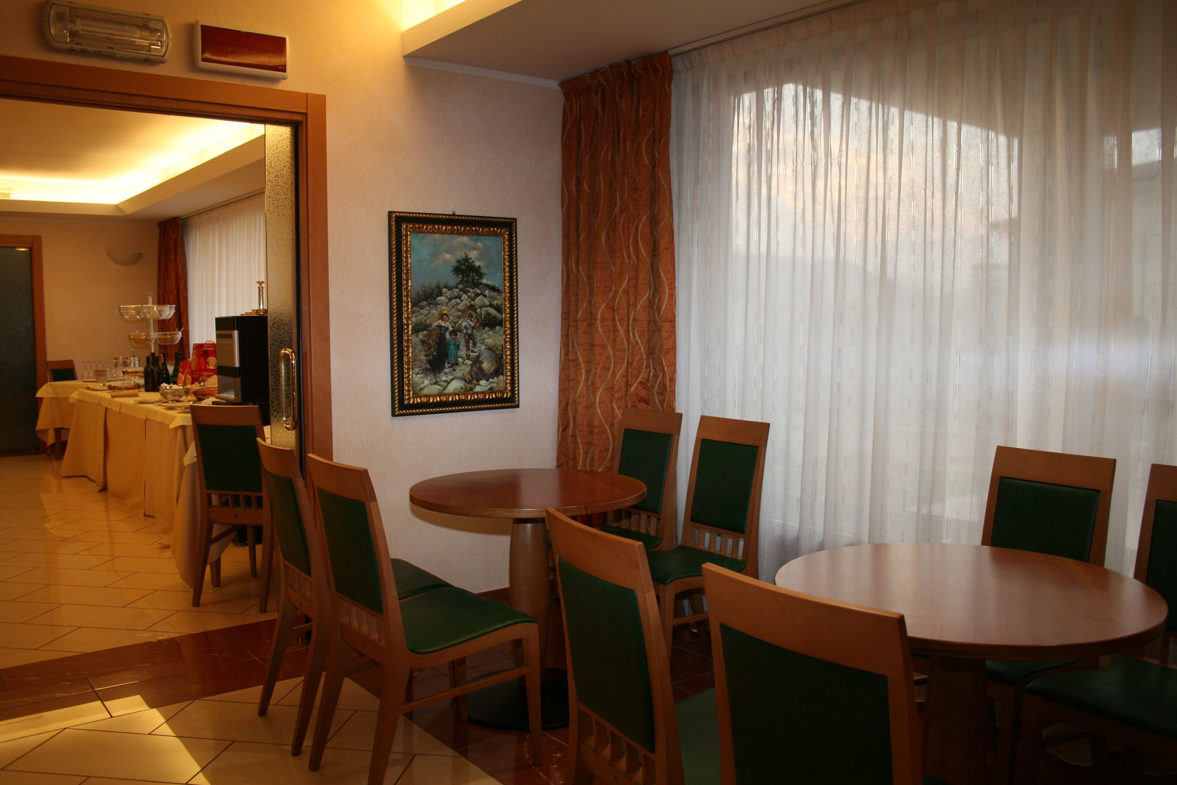 A dining area with round tables and chairs, a painting on the wall, and a view of the dining room through a doorway.