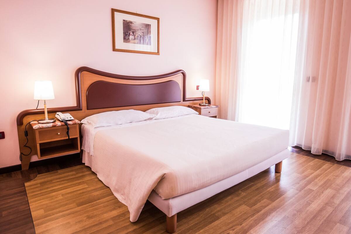 A neatly made bed in a hotel room with a wooden headboard, two bedside tables with lamps, a framed picture on the wall, and a large window with sheer curtains.