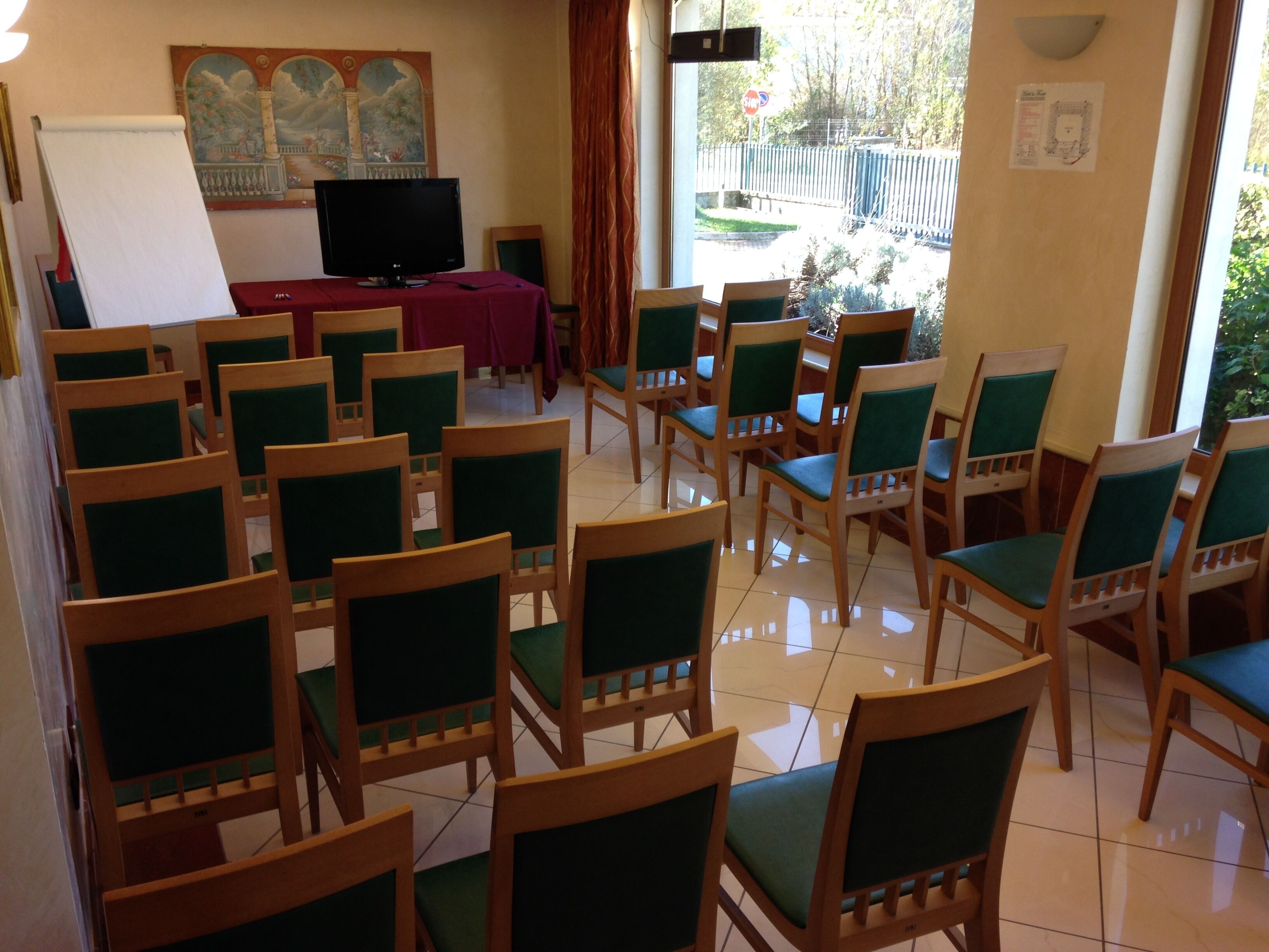 Une salle de conférence avec des rangées de chaises en bois dotées de coussins verts, un tableau blanc, un téléviseur et une fresque murale.