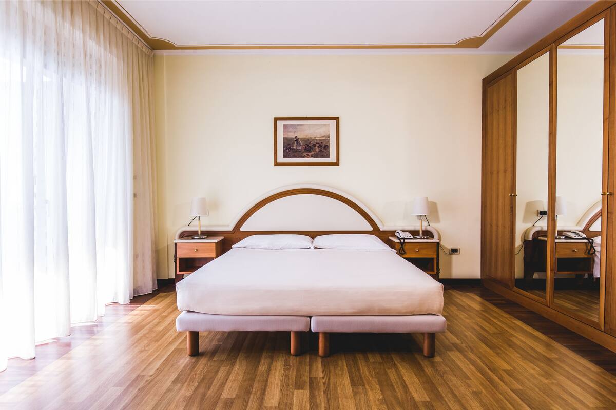 A hotel room with a large bed, wooden floor, a nightstand with a lamp, a bedside table with a lamp, and a large mirror cabinet.