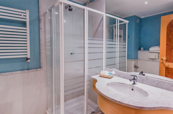 Un baño con ducha envuelta en vidrio, un lavamanos blanco con encimera de mármol y un espejo que refleja la habitación.
