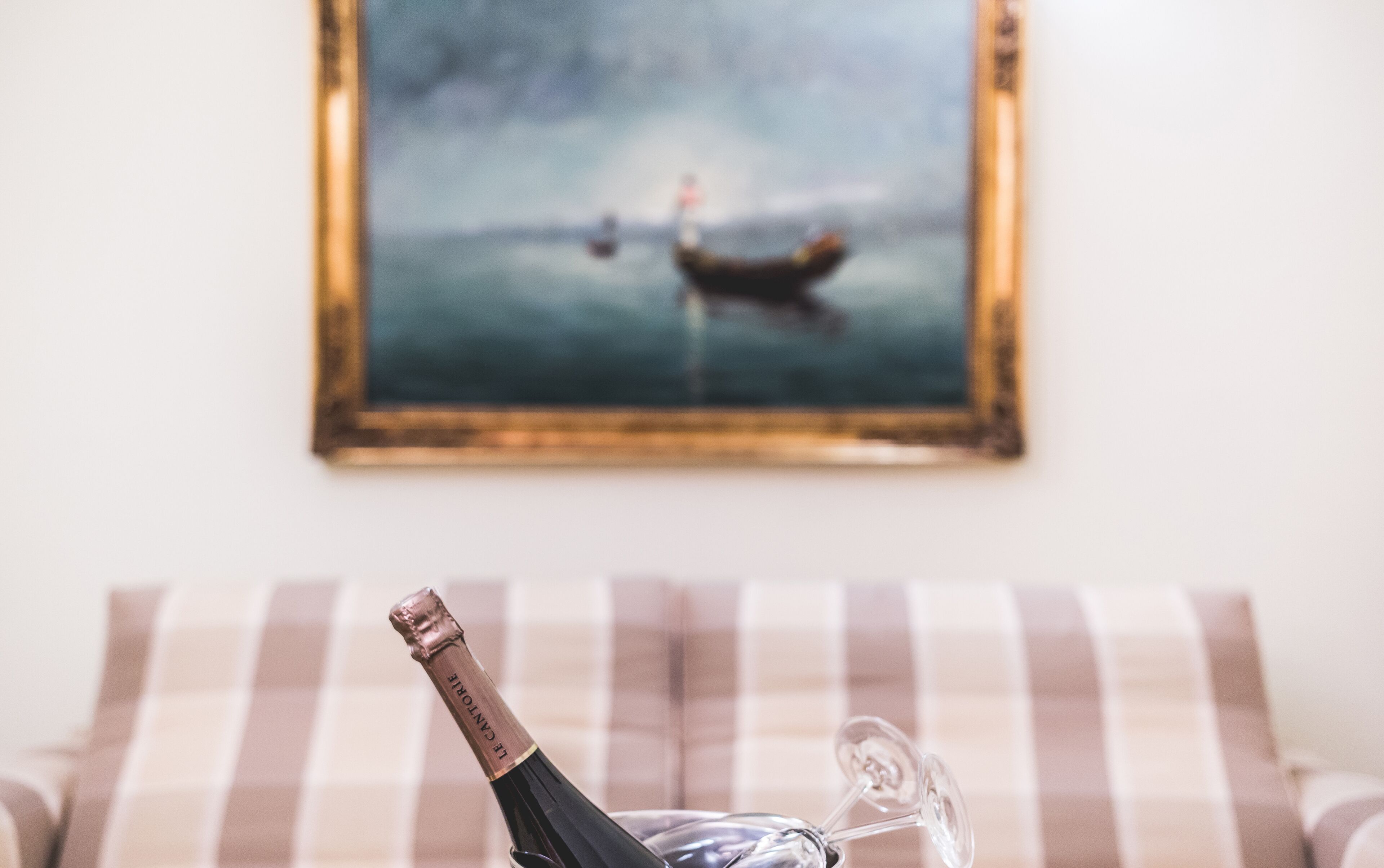 Une bouteille de champagne est posée sur une table. Au premier plan, on aperçoit un coussin rayé, et au fond, un tableau encadré représentant un bateau est accroché au mur.