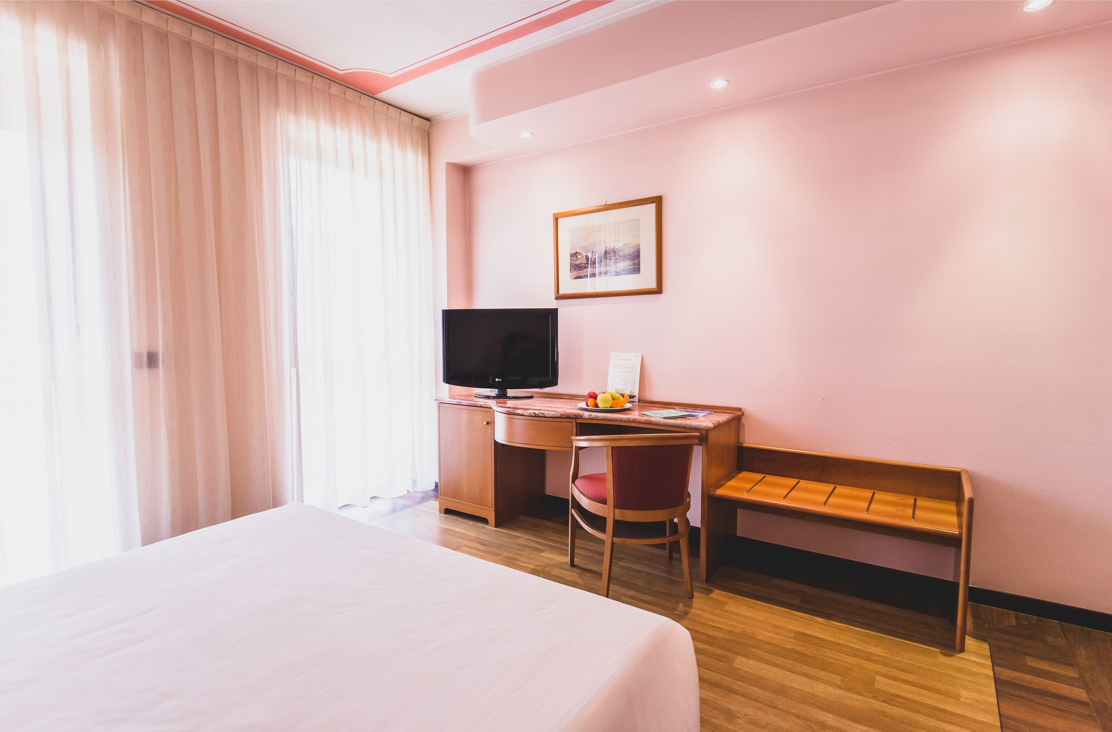 A hotel room with a bed, a desk with a computer, a TV, and a bench.