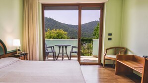 A hotel room with a bed, a desk with a lamp, a chair, a table, and a balcony with a view of a forested hill.