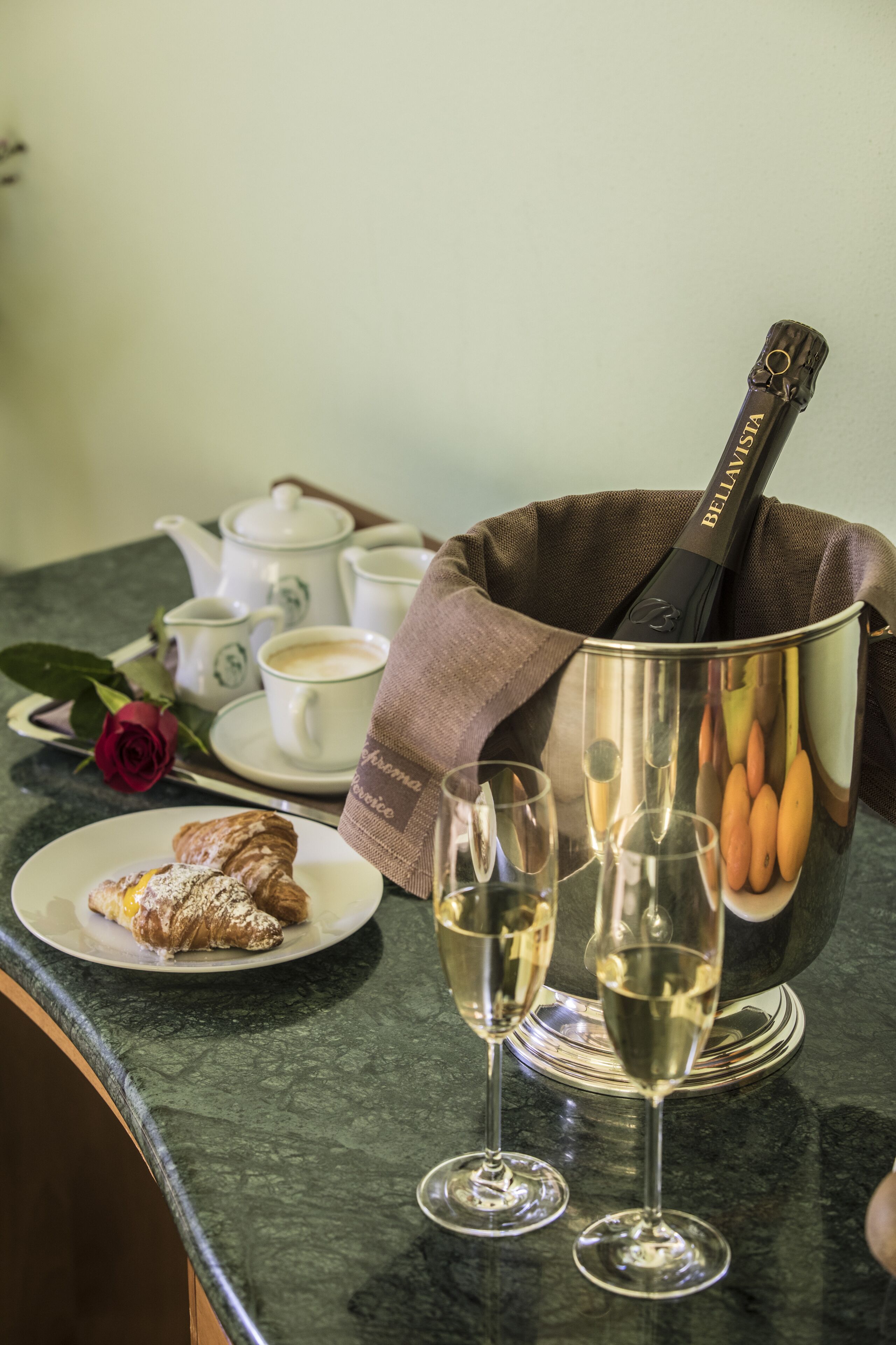 Une table avec une bouteille de champagne, deux verres, une théière, des tasses, une rose et une pâtisserie.