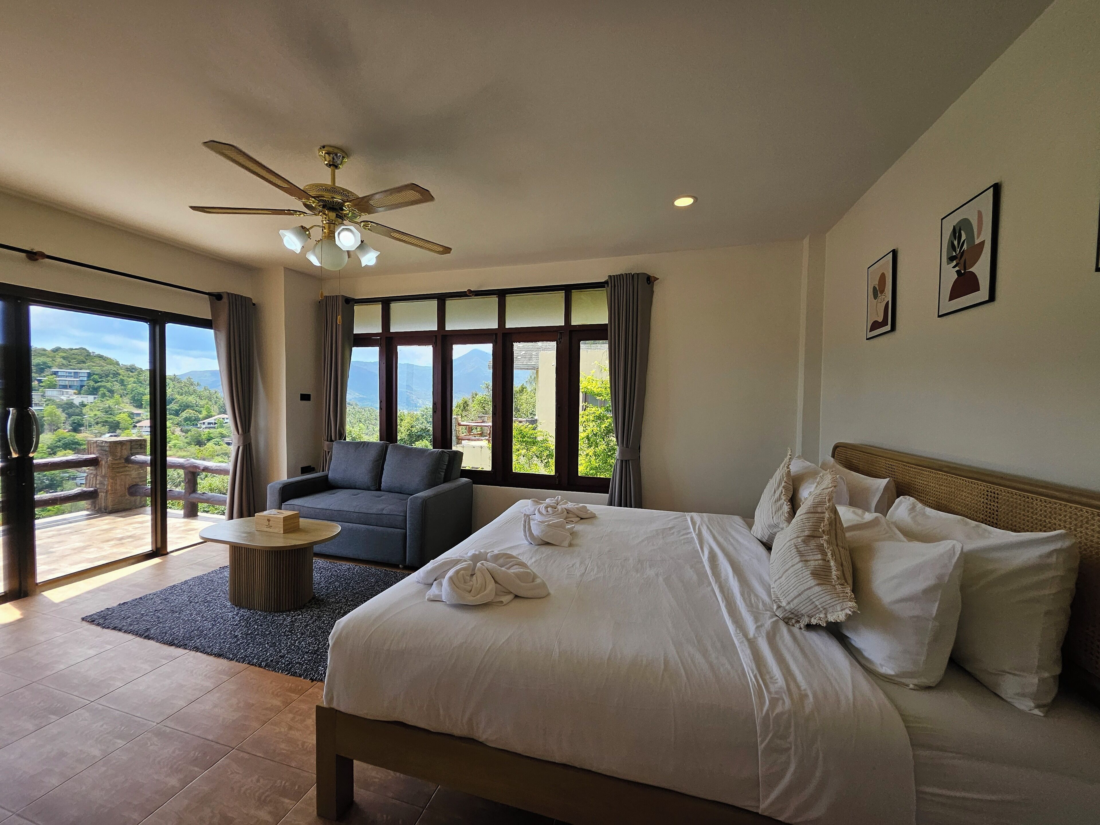 Quarto de hotel com cama, sofá, ventoinha no teto e grandes janelas com vista para as montanhas.