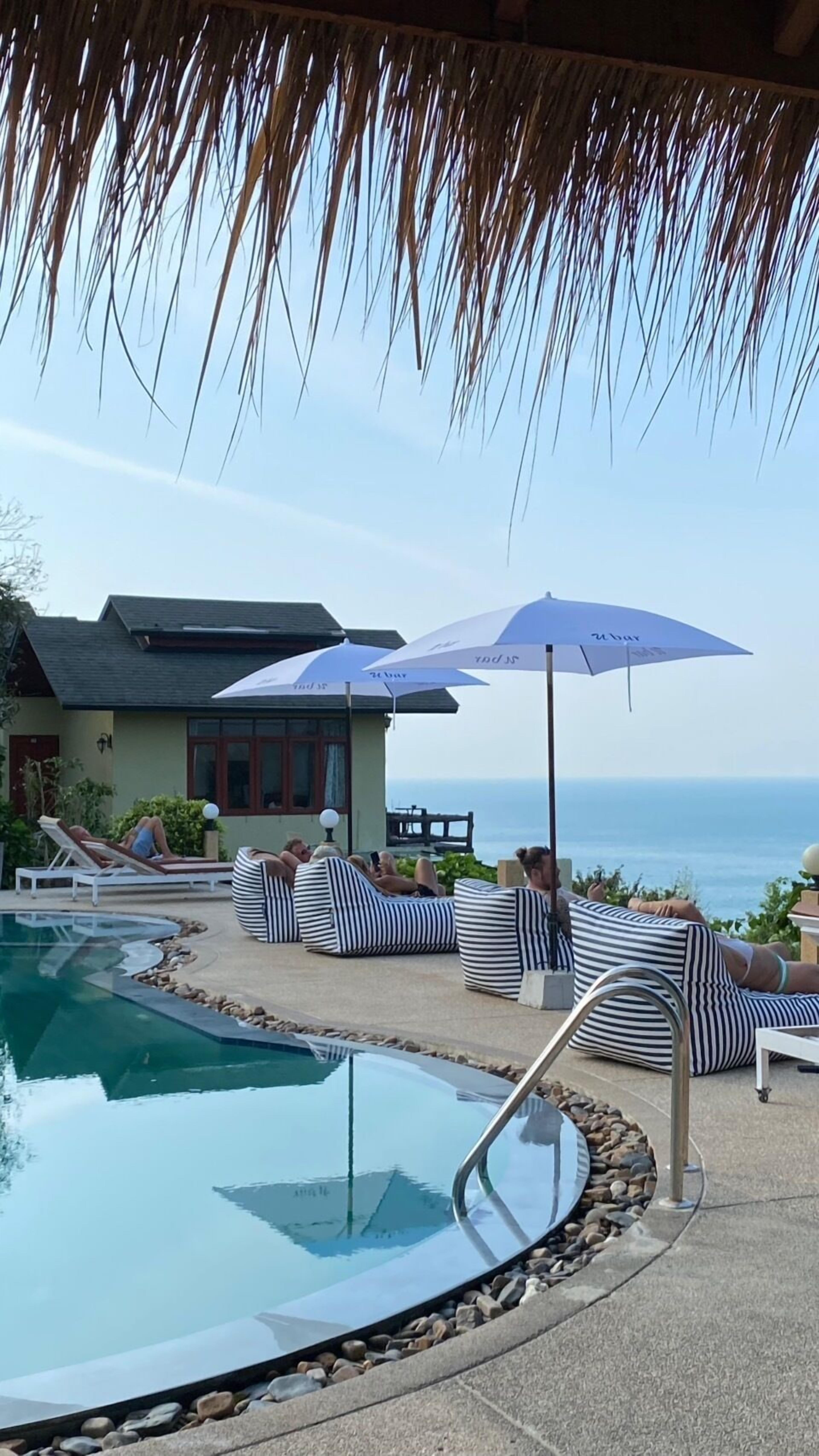 A pool area with lounge chairs and umbrellas, a house in the background, and a view of the ocean.