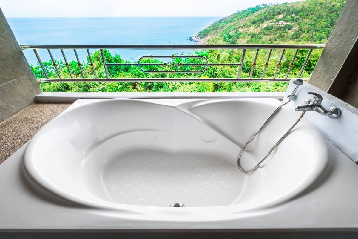 Una bañera blanca con cabezal de ducha, frente a un balcón con vistas al mar y al follaje.